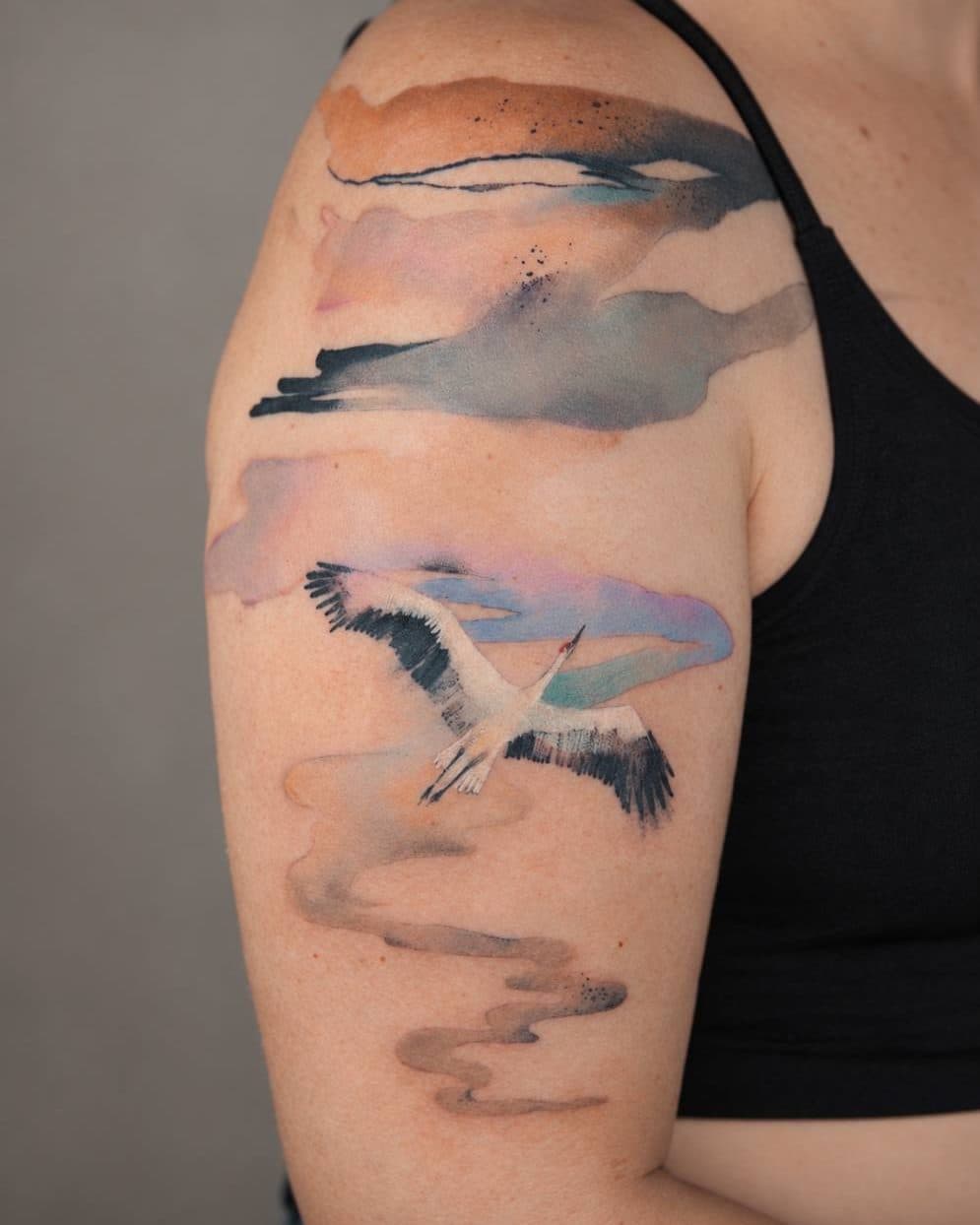 Watercolour tattoo of a crane flying through soft clouds, on an upper arm